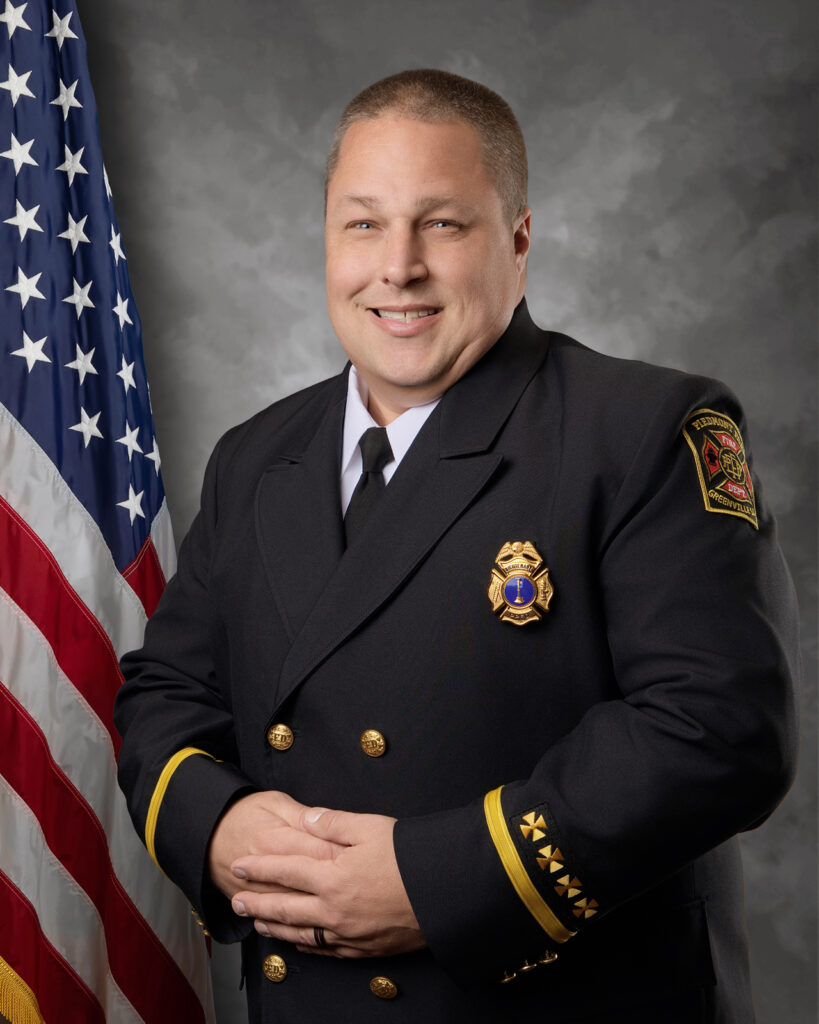 A man in a dark formal dress uniform with gold accents stands in front of an American flag, smiling, with his hands clasped. A fire department badge and patch are visible on his jacket.