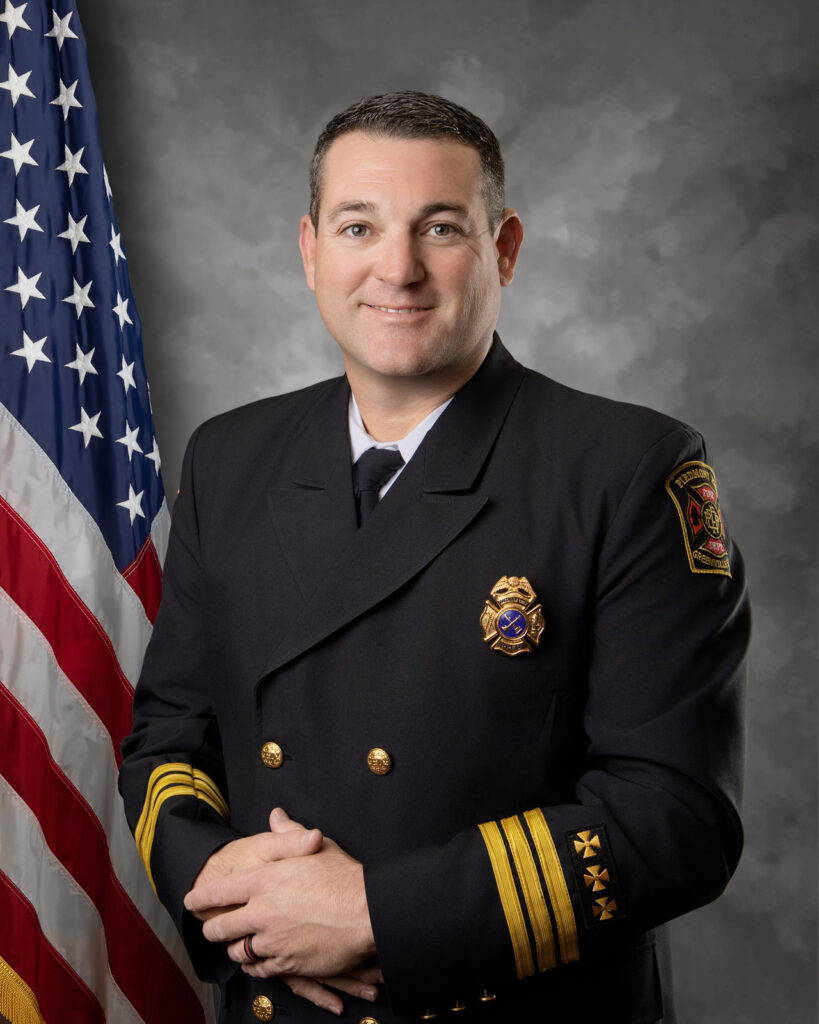 A man in a formal dark fire department uniform stands in front of a U.S. flag and gray backdrop, smiling with his hands folded. The uniform has gold buttons, insignia, and a badge on the chest.