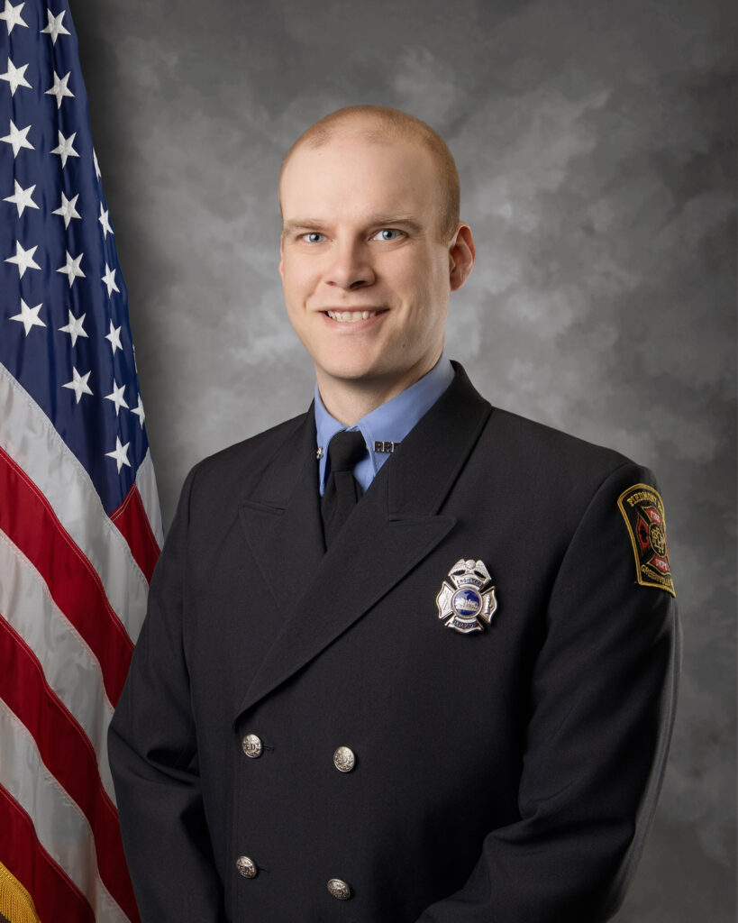 A smiling firefighter in formal uniform stands in front of an American flag and a gray backdrop. The firefighter is wearing a dark jacket with buttons, a badge, and a patch on the shoulder.