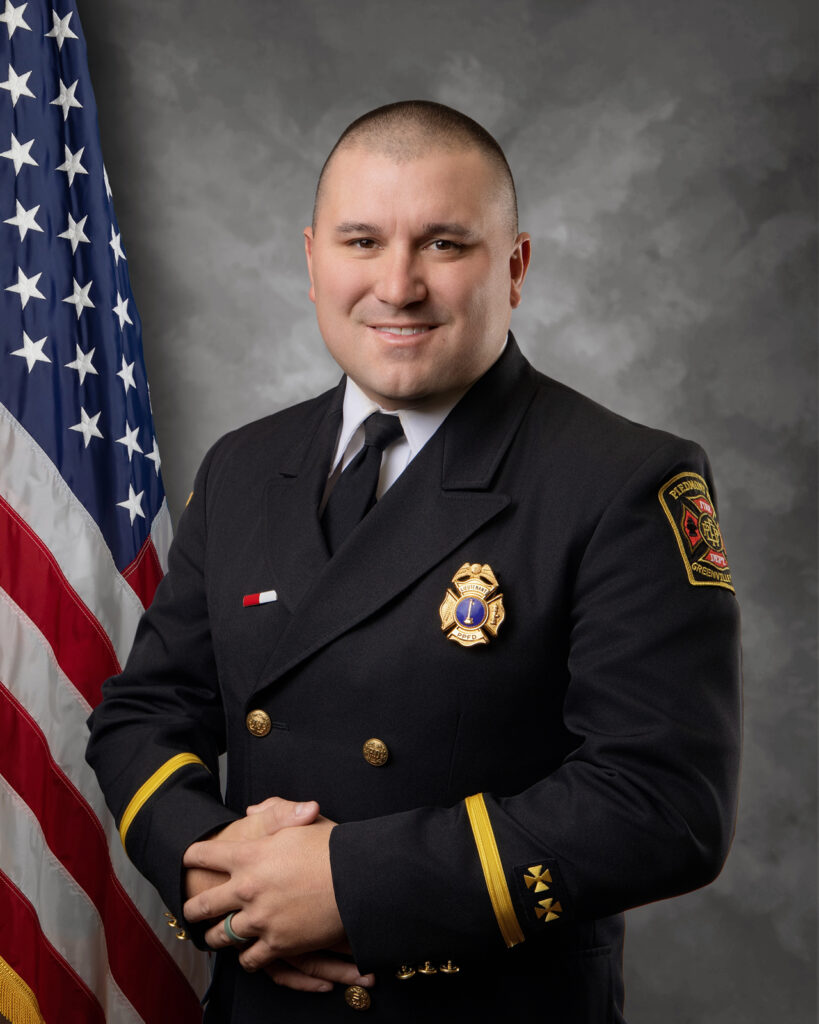 A man in a formal dark fire department uniform stands in front of a U.S. flag, smiling with hands folded. He wears a badge, insignia, and has short hair, against a gray cloudy backdrop.