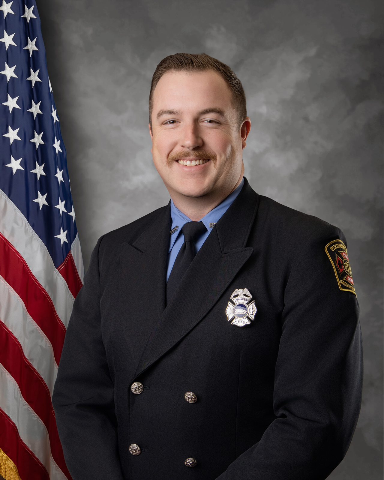 A firefighter in formal uniform stands smiling in front of an American flag and a gray backdrop. He has a mustache and short hair, and wears a badge and fire department patch on his jacket.