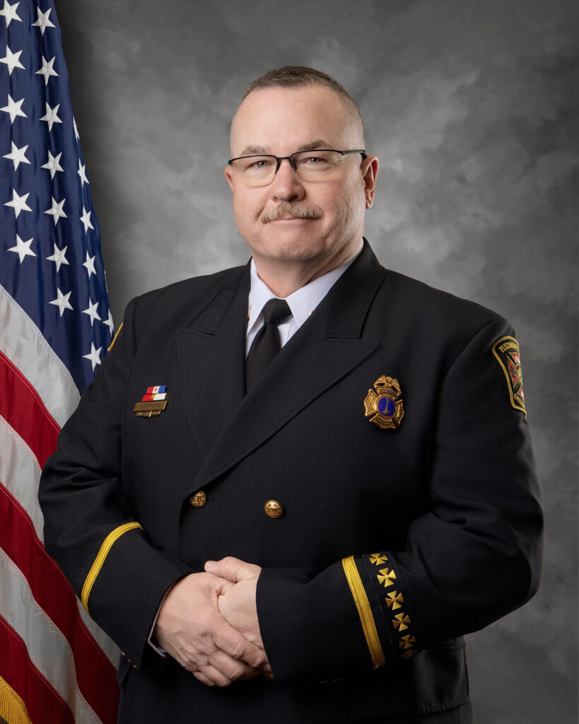 A man in a formal dark uniform with gold buttons and insignia stands in front of a US flag, hands clasped, wearing glasses, with a neutral expression and short hair.