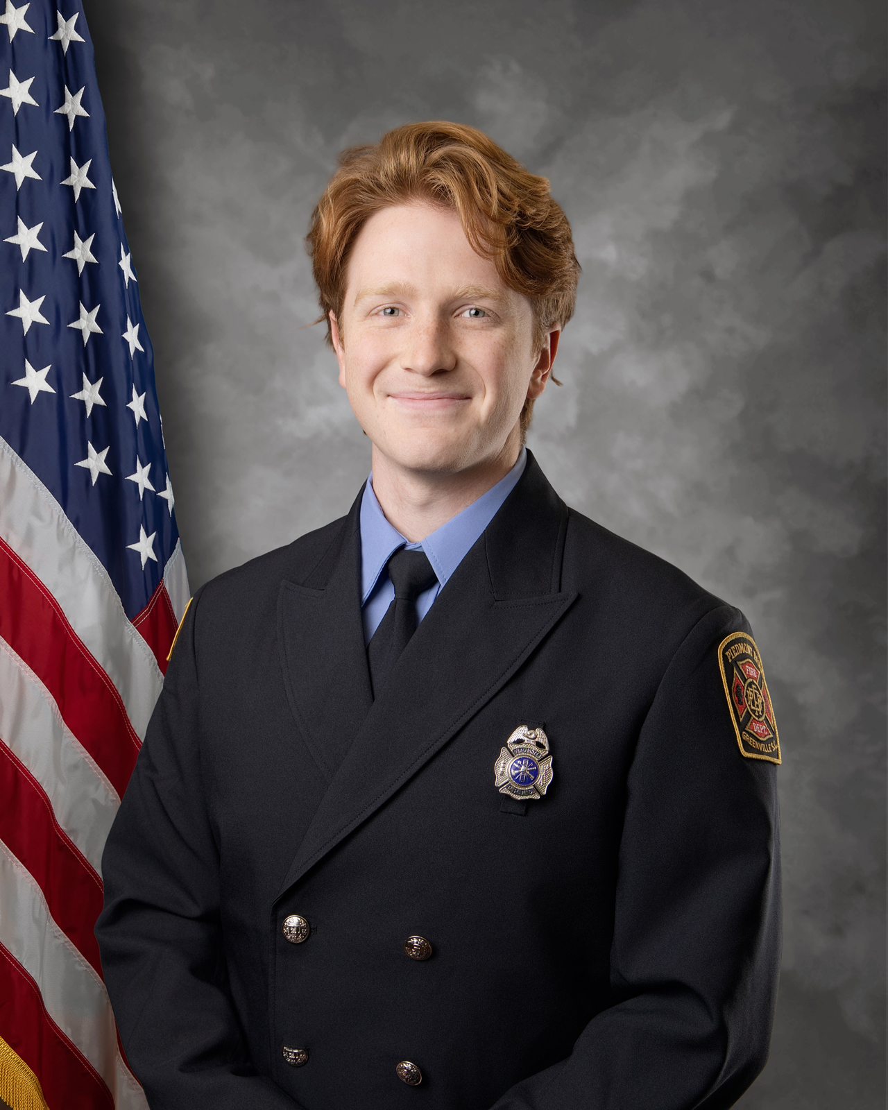 A firefighter in formal uniform stands in front of an American flag, smiling at the camera. The background is gray and the uniform features a badge and department patch on the arm.