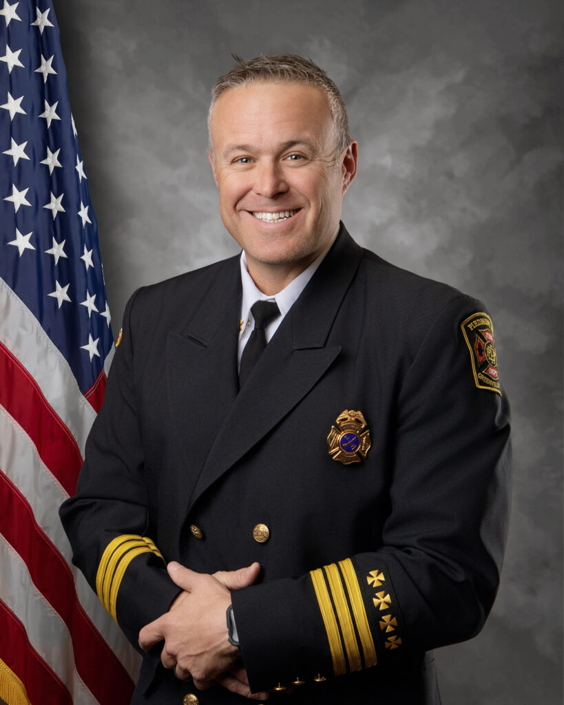 A smiling man in a formal fire chief uniform stands in front of a U.S. flag and a gray backdrop, with his hands clasped in front. His jacket has gold stripes and badges.