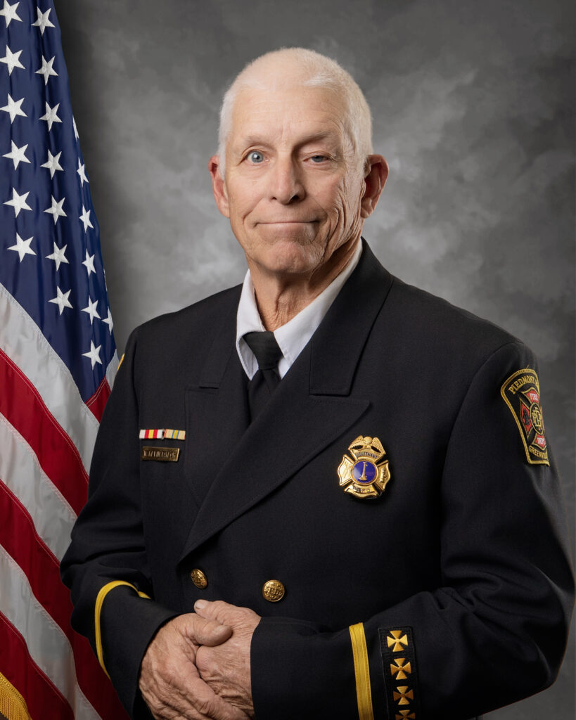 An older man in a formal fire department dress uniform stands in front of a U.S. flag and a gray backdrop. He has short gray hair, holds his hands together, and wears a badge, collar pins, and department patches.
