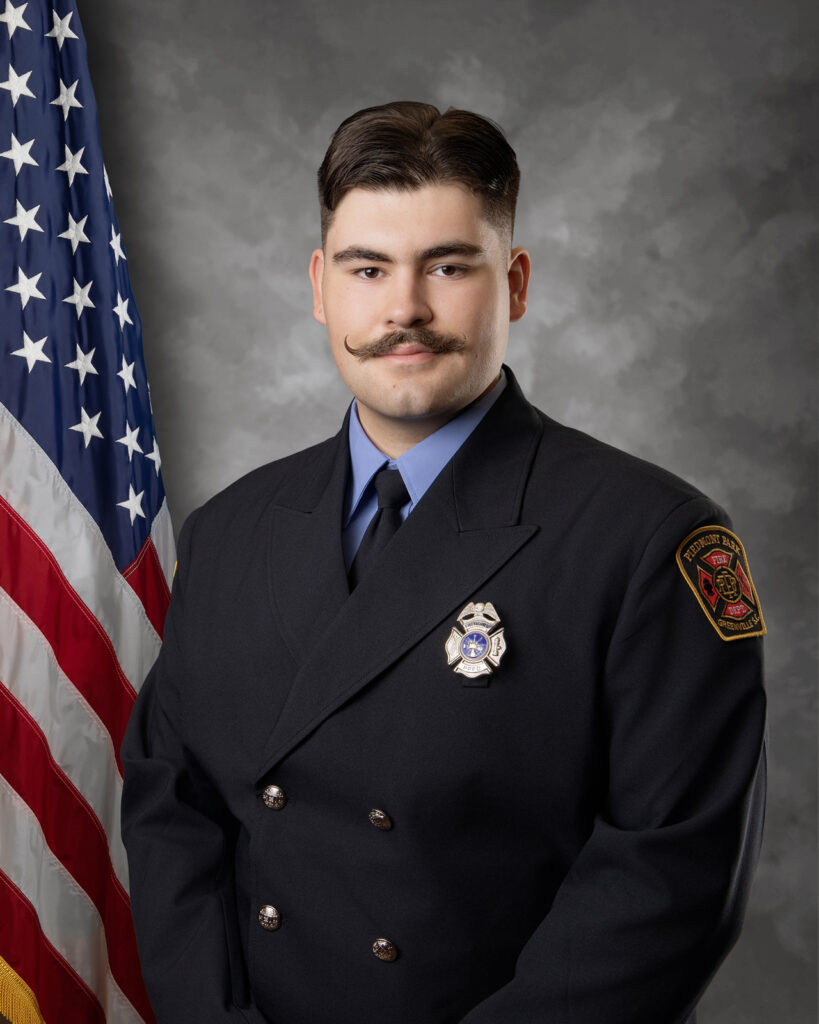 A firefighter with dark hair and a mustache wearing a formal uniform stands in front of an American flag and a grey backdrop, looking at the camera with a neutral expression.