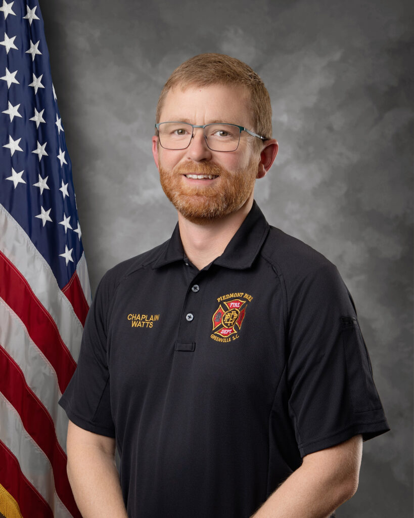 A man with short red hair, beard, and glasses stands smiling in front of a U.S. flag, wearing a black shirt with a fire department emblem and text that reads "Chaplain Watts.