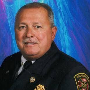 A man in a dark fire department uniform poses for a formal portrait against a vibrant blue and purple background. He has short gray hair, a mustache, and wears several badges on his jacket.